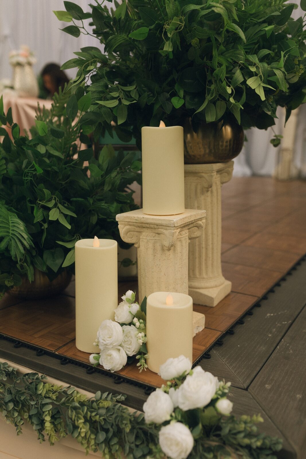 Pillar candles arranged on a column with cascading greenery and white roses at a reception backdrop.