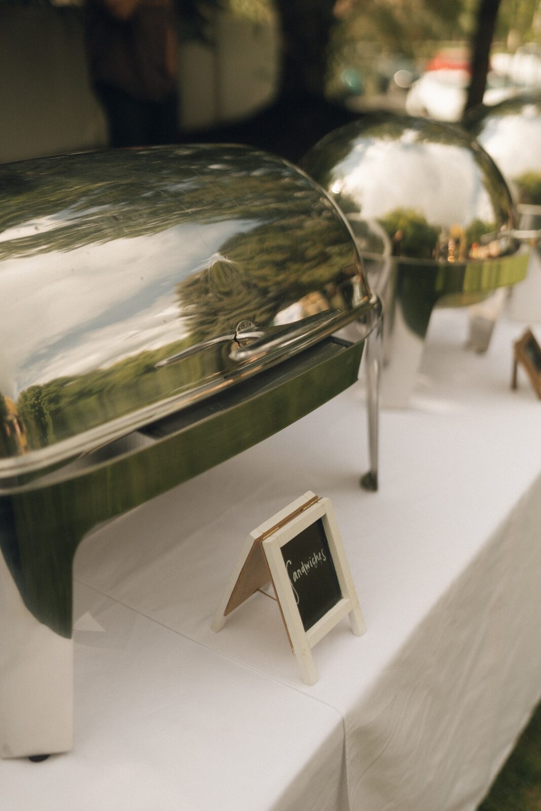 Polished silver chafing domes lined along a crisp white linen table with a hand-lettered chalkboard sign.