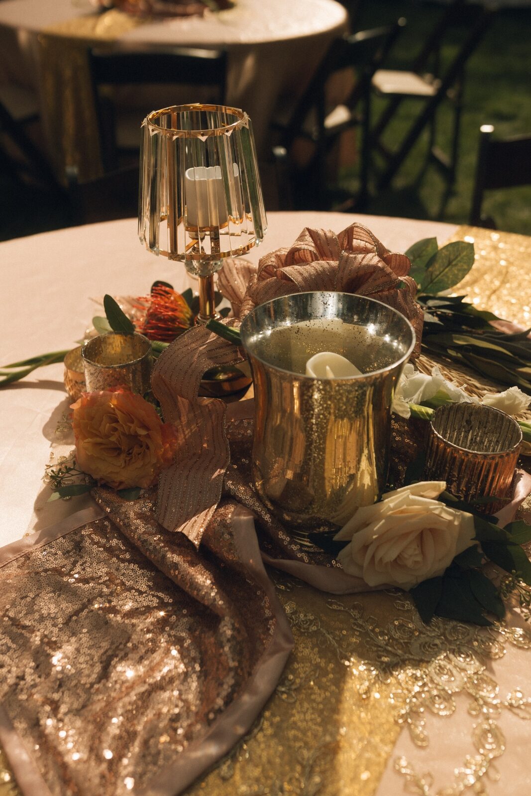Tablescape close-up with gold candle vessels, ribbon, sequined runner, and a single ivory rose.