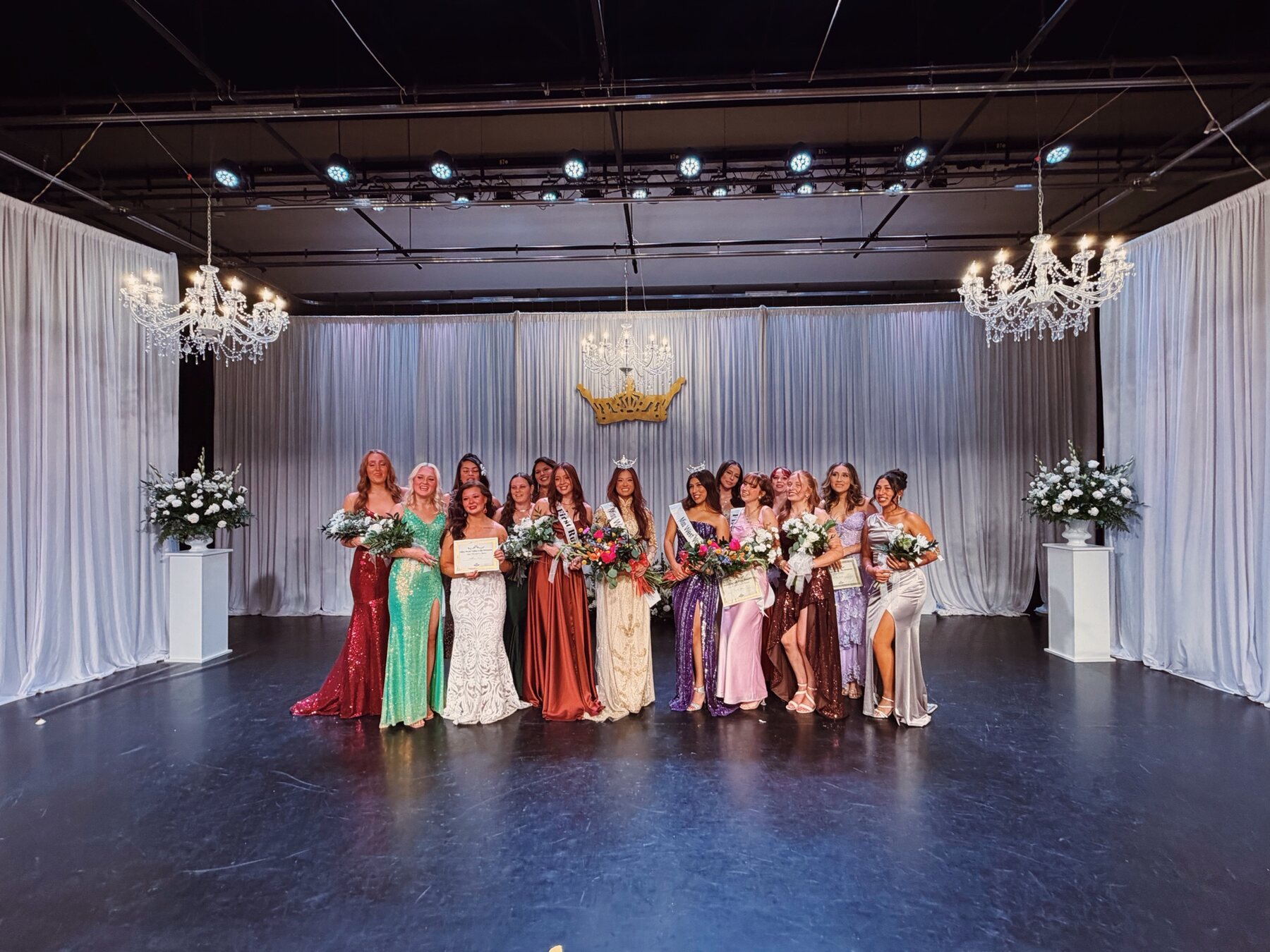 Group photo on stage at the Miss West Valley City event with white draping, chandeliers, and floral arrangements.