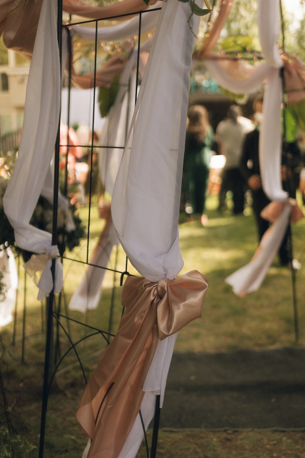 Flowing white and blush satin ribbons tied to an iron ceremony archway, lit by garden lights.